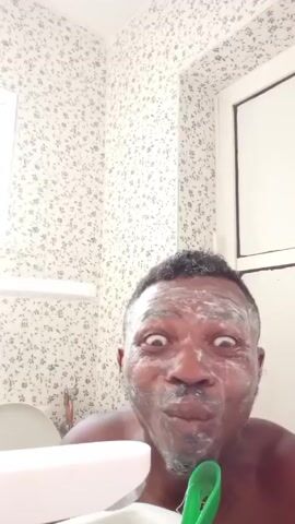 A close-up shot shows a dark-skinned man in a bathroom, his face covered in white cream. He has wide eyes and an open mouth, as if screaming. His hands are raised with fingers spread. He is shirtless, standing close to a sink, with a white container on the left. The bathroom has a floral wallpaper and a window in the background. The overall impression is startling and unusual.
