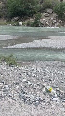 The video shows a natural scene with a river flowing through a rocky, mountainous landscape. The river has a greenish-blue hue and appears to be flowing over a shallow, gravelly bed, creating several smaller channels and sandbars. The banks of the river are covered in small stones and pebbles. In the foreground, a discarded plastic bottle lies on the gravel. Further into the river, a person, partially submerged and wearing light-colored clothing, is visible, seemingly interacting with the water. The background features steep, rocky slopes covered with trees and vegetation, with a hazy, possibly mountainous, vista in the distance. The lighting suggests it is daytime, possibly overcast or late afternoon.