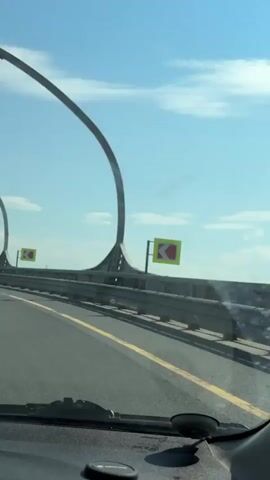 The video shows a car driving on a highway with distinctive curved lampposts. The road has yellow lane markings and a grey guardrail. A road sign with a left arrow is visible. Another car is ahead in the same lane. The sky is blue with scattered clouds. The view is from inside a moving vehicle.