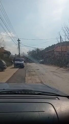 The view from inside a parked car shows a partially overcast sky over a rural street. Several vehicles are parked along the street. A building with a red tile roof sits behind a low stone wall. Bare tree branches reach upward to the sky. Power lines run along the roadside.