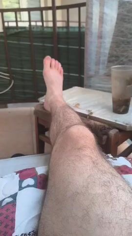 The video begins with a close-up of a person's bare foot and leg, which is hairy. The person's foot is resting on a wooden stool. In the background, there is a glass with a dark liquid inside, and what appears to be a chair covered in plastic wrap. The scene then pans out to reveal a balcony of an apartment building. There is a railing in the foreground, and a green fence behind it. Further in the background, there are other balconies on the apartment building, and a blue and green umbrella. The camera continues to pan up, showing more of the building and the sky. The video appears to be taken on a clear day.
