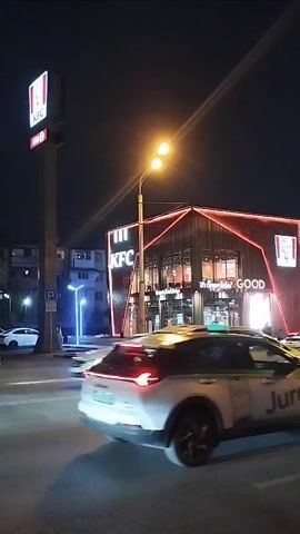 This video shows a nighttime scene at a KFC restaurant in Dushanbe, Tajikistan. The KFC building is modern and has prominent red neon lighting outlining its angular roof. The KFC logo is clearly visible on the building and on a tall sign. The text "GOOD" is also visible on the building, likely part of a slogan or a separate business.

The setting is an urban intersection with several cars driving by. One car in the foreground is a white SUV with its taillights illuminated. Another vehicle, a white car with green accents and a taxi sign on its roof, is also visible. The streetlights are on, casting a warm glow on the scene. The sky is dark, indicating it is nighttime. There is no visible weather, but the lighting suggests clear conditions. No people are clearly discernible due to the low light and motion blur. The overall mood is that of a typical evening in a city, with the bustling activity of traffic and a popular fast-food restaurant.