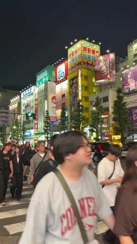This video captures a night scene on a busy street, likely in Japan, given the signage. A crowd of people is crossing a crosswalk. The buildings on either side are brightly lit with large neon signs advertising various businesses. One prominent sign, in yellow and red, features Japanese characters and images of anime-style characters. Another green sign reads "GIGO." Streetlights are visible, and trees line the sidewalk. People are dressed in casual modern clothing. Some individuals have backpacks. The overall atmosphere suggests a lively urban environment, possibly a popular entertainment or shopping district.