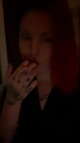 A woman with red hair and tattoos is smoking a cigarette. She takes a drag and exhales a large cloud of smoke into the camera. She is wearing a black shirt. The background is dark and indistinct.