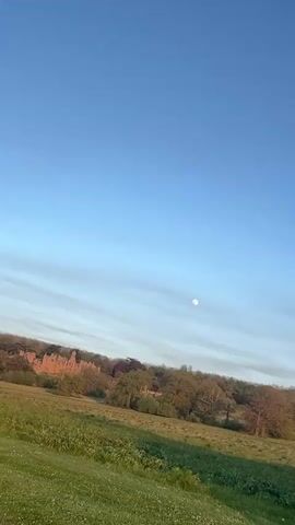 This video features several clips of an evening landscape in South Kesteven, United Kingdom. The time of day appears to be late afternoon to sunset, indicated by the warm hues in the sky during later clips.

The first clip shows a wide shot of a vast green field with scattered trees and distant structures under a clear blue sky, with a visible full or near-full moon centered in the frame. The sky transitions from a bright blue at the top to a lighter, more hazy blue-white near the horizon.

The second clip is a close-up of lush, dark green hedges against a bright blue sky, suggesting a well-maintained natural boundary.

The third clip reveals a paved pathway running alongside a wire fence and a green hedge. A bare tree with emerging leaves stands prominent against the sky, which now displays soft orange and yellow tones of a sunset. Distant figures can be seen walking on the path.

The final clip provides a broader view of the pathway winding through expansive green fields and past clusters of trees. The sunset colors are more pronounced, painting the sky with serene oranges and yellows. In the distance, several houses or buildings are visible, nestled among the trees.

The overall scene depicts a tranquil, open rural or semi-rural environment during a beautiful sunset, with no discernible text or specific human activity beyond implied observation or walking. The weather appears clear and calm throughout.