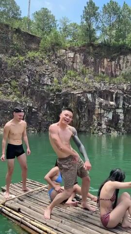 A group of four people are on a bamboo raft in a quarry with green water. On the raft, there are three people visible, two men and one woman. One man, wearing black swim trunks, stands at the back of the raft. Another man, wearing patterned shorts, is in the center of the raft, dancing. He has tattoos on his left arm. A woman in a bikini is seated on the raft near the man in shorts. In the water, there is a third person, a woman wearing a bikini, swimming. The quarry walls are steep and rocky, with some trees and vegetation growing on them. The sky is clear and blue.