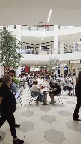 The video shows a bustling food court in a multi-story shopping mall. In the foreground, two people are walking away from the camera. One is wearing a black t-shirt and dark pants, the other is wearing a black t-shirt and black pants. They are walking across a patterned floor made of hexagonal tiles in shades of gray and white. In the midground, several people are seated at round tables in the food court, eating and socializing. Behind them are various food stalls with signage, including "ZALES." The upper levels of the mall are visible, with more shops and balconies overlooking the food court. Trees are placed around the seating area, and overhead lighting illuminates the space. The overall atmosphere is one of a busy public space.