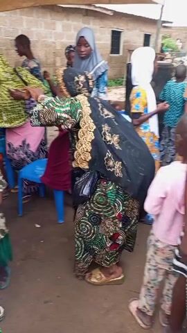 Here is a description of the video:

The video shows a group of people, primarily women, gathered outdoors. Most of the women are wearing traditional or colorful dresses and head coverings. One woman stands out because she is wearing a heavy black garment adorned with gold floral embroidery, seemingly being adjusted or supported by another woman. A blue plastic chair is positioned near her. Other women and children are standing nearby, some of them watching the scene. A man in a brown outfit walks by in the background. The setting appears to be a courtyard or an open area with simple buildings visible in the background. A temporary shelter seems to be provided by a structure made with poles and a fabric or plastic roof.
