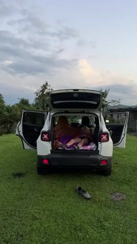 The video shows the rear of a white Jeep with its hatchback and rear doors open, parked on a grassy area. Inside the cargo area, two people are visible. One person, covered by a brown blanket, is lying down. Another person, with blonde hair and tattoos on their legs and arms, is lying on their back on a pink and white patterned mattress, covered by a purple sleeping bag. This person is wearing a pink shirt, black shorts, and patterned socks. Their head is propped up on a pillow. A pair of black clogs are on the ground in front of the car. The background consists of green foliage and trees, with a cloudy sky overhead. The video progresses with a zoom effect, focusing more closely on the person lying in the cargo area.
