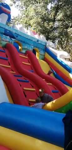 The video shows a close-up, slightly shaky shot of a colorful inflatable slide. Two young boys are climbing the slide. The boy in front is wearing a gray t-shirt and shorts and has blonde hair. The boy behind him is wearing a light blue shirt and white shorts. They are both gripping the red climbing holds. The inflatable structure is decorated with blue, white, red, yellow, and green colors, and appears to be set up outdoors with trees visible in the background. The lighting suggests it is daytime.