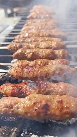 A close-up, slightly tilted shot captures a row of kebabs on a grill. The kebabs are elongated and appear to be made of ground meat, skewered on metal rods. They are positioned horizontally across a metal grill grate, with charcoal visible beneath. Smoke rises from the grill, obscuring some of the kebabs further back. The foreground shows the charring and browning of the meat, indicating it is cooking. The lighting suggests an outdoor setting, possibly during the day, with hints of green foliage in the blurred background. The camera focuses on the kebabs, with a shallow depth of field blurring the background and foreground elements.