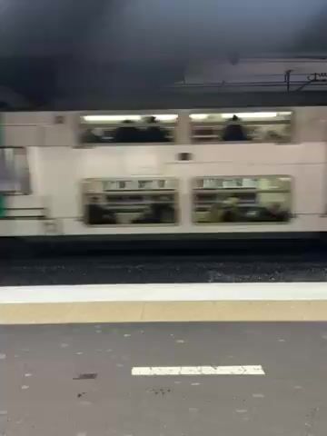 The video shows a double-decker train moving past the camera at a subway station. The train is white with a green stripe, and passengers can be seen through the windows. The subway platform has a gray surface with a yellow and white striped safety border. A person walks in the background of the frame.