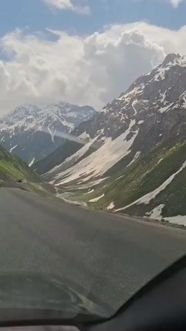 The video shows a view from inside a vehicle driving on a paved road through a mountain pass. The mountains are partially covered in snow with green vegetation on the lower slopes. The sky is blue with white clouds. There is a truck driving in the opposite direction on the road. The windshield of the vehicle is visible. The lighting is bright and sunny.