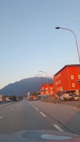 The video shows a forward-facing view from inside a vehicle traveling on a road. To the right of the road are two red buildings with multiple floors and many windows, appearing to be commercial or residential structures. A street light is visible between the buildings. Further down the road, a tall sign with text and an abstract design stands on the right. Mountains are visible in the background, with a clear blue sky above, suggesting it is either sunrise or sunset due to the warm lighting on the buildings. Several cars and a van are parked or driving on the road and in the parking areas next to the buildings. The road has white lane markings and a barrier on the left side. The overall scene depicts a town or suburban area with a natural mountainous backdrop.