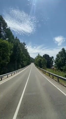 The video shows a first-person view of a car driving on a paved road with a white dividing line. The road is surrounded by lush green trees on both sides, with a clear blue sky above and some scattered white clouds. A metal guardrail runs along the side of the road. The camera pans slightly, showing the interior of the car on the right side of the frame. The sun is shining brightly, casting shadows.
