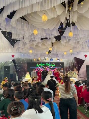 The video captures a wedding reception taking place inside a large tent, which is decorated with white fabric draping from the ceiling, resembling clouds, and interspersed with decorative lights. The focus is on the newly married couple standing on a raised stage, flanked by family members. The bride wears a traditional white gown, while the groom is in a formal suit. A large LED screen behind them displays a message in Vietnamese. A multi-tiered wedding cake is visible on the stage as well.

The audience, seated at tables, are dressed in a variety of attire and are observing the proceedings, with some people holding up their phones to take photos or videos. A photographer, identifiable by his camera equipment, is positioned in front of the stage, capturing the scene. Sparklers are being set off on either side of the stage, adding to the celebratory atmosphere. The tent is filled with guests and the general atmosphere appears festive.