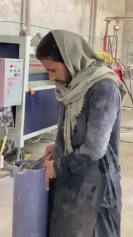 Here is a detailed description of the video:
A man stands indoors, operating equipment. He is light-skinned with dark hair and a trimmed beard. He wears a grey headscarf and a dark, paint-splattered overall. He uses a tool, possibly a wrench, to interact with the interior of a large grey plastic pipe, which he holds with his left hand. Behind him is a machine with a blue and white color scheme, featuring indicator lights on a control panel. Yellow cables and hoses are visible connecting to the machine. In the background are blurred elements, including what appears to be a scaffolding-like structure and additional machinery. The environment appears to be a manufacturing or workshop setting.