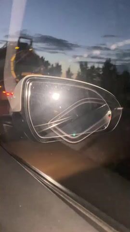 The video shows a close-up view of a car's side-view mirror at dusk or night. The mirror reflects the lights of other vehicles on a highway and the dark outline of trees and hills in the background. The camera appears to be inside the car, looking out of the window at the mirror. The reflection in the mirror shows bright lights from headlights and taillights, suggesting traffic. The sky is dark blue, indicating the end of daylight. There are no people clearly visible in the video, but the presence of the car and other vehicles implies human activity. The environment is a road at night, likely a highway due to the presence of multiple lanes of traffic indicated by the lights.