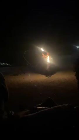 At nighttime, a person performing a fire show is seen engulfed in a large ball of flame. They are holding a burning object in their hand, possibly a torch. In the background, a metal structure in the shape of a heart is illuminated by the fire. The ground is covered in what appears to be gravel or small stones. Two other people are visible in the foreground, sitting and observing the performance. The overall scene is dark with the bright flames of the fire show being the primary source of light.