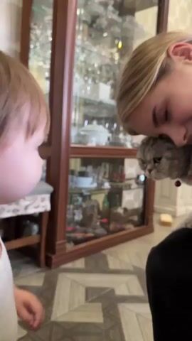 A young child with brown hair is facing right, wearing a white tank top. The child's hands are touching a gray cat's head. To the right of the child, a young woman with blonde hair is leaning in, holding the cat and smiling. The woman is wearing black clothing. They are indoors, in what appears to be a living room with a display cabinet in the background. The floor has a patterned tile design.