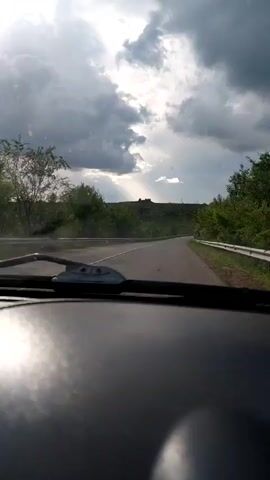 The video captures a first-person perspective from inside a moving vehicle, looking forward through the windshield. The foreground shows the car's dark dashboard reflecting the sky, with a windshield wiper base prominently visible.

The scene depicts a paved road, appearing to be a two-lane highway, gently curving ahead and slightly uphill. Green trees and dense vegetation line both sides of the road. On the right side, a metal guardrail runs alongside the road. In the distance, rolling hills covered in greenery are visible under a dynamic sky.

The sky is heavily overcast with large, dark gray clouds, but bright shafts of sunlight break through, creating a dramatic lighting effect on the landscape. This suggests a partially cloudy day, possibly indicating variable weather conditions.

A red circular traffic sign with a diagonal line (indicating a prohibition, likely "No Stopping" or "No Parking") is visible on the right side of the road. Below the red circle, there is text in Cyrillic, which appears to read "250 м," signifying a distance of 250 meters.

The activity shown is driving along this rural or semi-rural road in the Agapovka area of Russia.