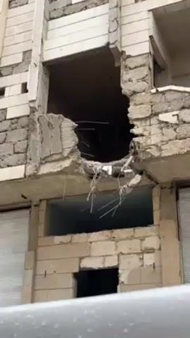 The video displays a close-up of a severely damaged building facade. A large, gaping hole penetrates a section of the upper floors, revealing exposed rebar and rubble. The lower level features shattered windows and missing bricks, indicating widespread structural damage. The building appears to be constructed from concrete blocks and stone. The footage is shot from a low angle, possibly from a vehicle passing by. The overall impression is one of destruction and devastation.
