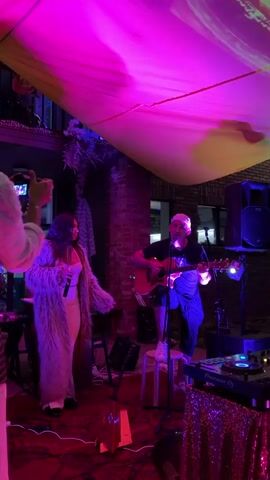 A live music performance is taking place, illuminated by vibrant pink and yellow lighting that reflects off a draped canopy overhead. In the foreground, a woman stands on the left, wearing a long, fuzzy, light-colored coat over a peach-colored outfit and holding a flute to her lips. She is facing a man on the right, who is seated on a white stool and playing an acoustic guitar. He is wearing a dark t-shirt, a light-colored cap, and dark shorts. Behind them, a brick wall forms the backdrop. To the left of the woman, musical equipment, including a keyboard and stands, is visible. To the right of the man, a DJ setup with turntables and a speaker are present. The floor is covered with a red rug. The overall scene suggests an indoor or semi-outdoor venue, possibly a bar or event space, with a festive atmosphere.