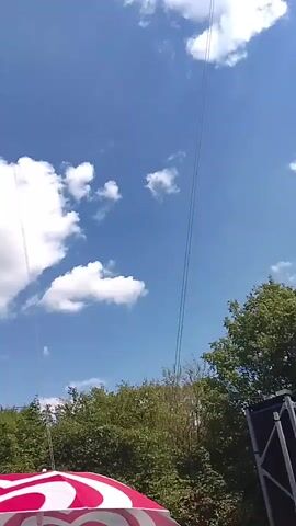 This media file captures a thrilling activity under a bright, blue sky dotted with fluffy white clouds, suggesting a clear, sunny day. The scene appears to be an outdoor amusement park or adventure facility, hinted at by the presence of a tall, lattice tower on the left, likely part of a ride.

The primary subject of the video is a high-speed launch of a human slingshot ride. At the beginning of the clip, a red and white object, possibly part of the ride's structure or a safety net, is visible at the bottom of the frame. The camera then pans up to reveal the clear blue sky.

The main event occurs around the 00:01 mark, where a circular cage with people inside is launched upwards with immense speed. This cage is attached to elastic cords that are anchored to the tall tower. The individuals inside are momentarily seen suspended high in the air before their trajectory begins to arc. The excitement is palpable, with a person heard exclaiming "Oh my God" and another shouting "Whoa!" at different points in the recording, indicating surprise and exhilaration.

The setting is a natural landscape, with dense green trees forming a backdrop below the sky and the ride. The tower and the launch mechanism are the prominent man-made structures in view. The activity is a bungee-like launch, designed to propel individuals to significant heights at high velocity. The video's perspective is from the ground, looking upwards, emphasizing the scale and speed of the launch. No visible text is present in the media. The location context provided is Rammingen, Germany, which further grounds the scene to a specific geographical place.