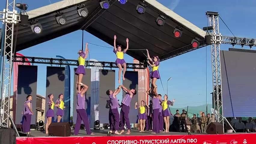 A group of performers, clad in purple and yellow costumes, are showcasing an acrobatic routine on an outdoor stage. The male performers, in purple pants and vests, are supporting female performers in purple skirts and yellow tops on their shoulders. One female performer executes a flip in the air. The stage is covered by a roof-like structure with lighting fixtures. A banner with Russian text is on the front of the stage. Behind the stage are video screens, and a crowd of people is visible in the background. The sky is blue and sunny.