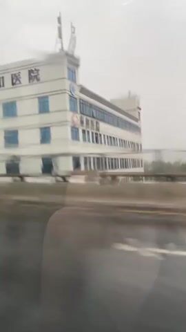 The video shows a side view of a multi-story white building with blue windows, identified by Chinese characters on its facade as "泰和医院" which translates to "Taihe Hospital." There are also signs indicating it is a rehabilitation center. The building appears to be located near a road or highway, as indicated by the blurred motion of a barrier in the foreground and the appearance of a roadway. The sky is overcast and appears to be raining, with reflections visible on the road surface. The building has antennas on its roof. The overall scene suggests the footage was captured from a moving vehicle.