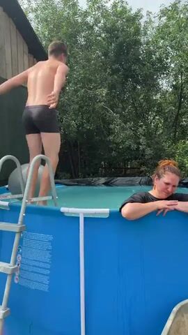 A young man in black swim trunks climbs out of an inflatable swimming pool and jumps back in, creating a large splash. A woman with her hair pulled back in a bun leans on the edge of the pool, watching him with a smile. The pool is blue and surrounded by lush green trees and a wooden structure. The sky is overcast.