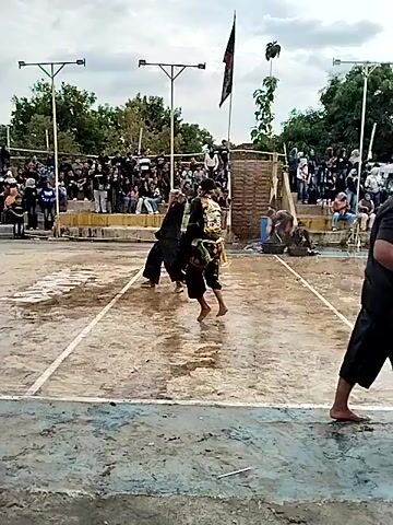 The video depicts an outdoor event, possibly a cultural performance or competition, with a crowd gathered to watch. Two individuals, dressed in what appear to be traditional costumes, are the central focus of the action. One performer, wearing a dark outfit with a distinctive headdress that resembles a bird or animal, is in motion. The other performer, in lighter, patterned attire, is facing away from the camera and seems to be in a defensive or responsive stance. The ground is wet, suggesting recent rain or the use of water, and there are some bamboo-like structures and flags in the background. The crowd is dense, with many spectators standing behind a barrier. The overall atmosphere appears lively and engaged.