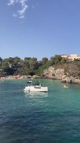 A drone-like view pans over a crystal-clear turquoise bay filled with people enjoying the water. The bay is surrounded by lush green trees, rocky cliffs, and buildings perched on the hillsides. Several boats, including a large white yacht with people lounging on it and a smaller speed boat, are anchored in the water. Numerous individuals are swimming, paddleboarding, and relaxing on inflatable tubes in the shallow, transparent water. The scene is bathed in bright sunlight under a clear blue sky.