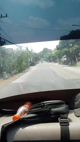 The video shows a view from inside a vehicle, looking out through the windshield. A road stretches ahead, lined with trees and buildings. The sky is visible through the windshield, with clouds and power lines. On the right side of the frame, a black bag and an orange-capped bottle of water are visible. The vehicle appears to be moving, as indicated by the slight blur of the passing scenery. There are a few cars and motorcycles on the road, as well as some people walking along the side. The overall impression is of a typical scene in a rural or suburban area.