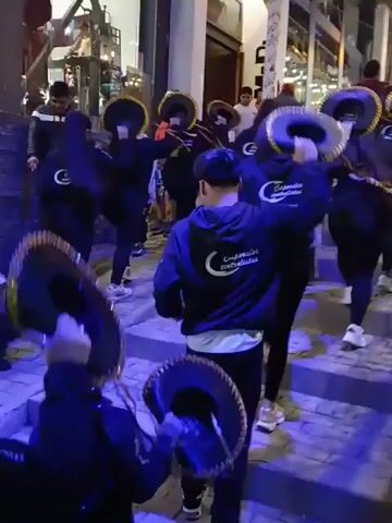 A group of people walks down a pedestrian walkway. Some wear matching dark-blue hoodies with white designs, and carry tambourines. The group walks past a cafe. People carry bags and other items on a wheeled cart. The street is illuminated by blue and yellow light.