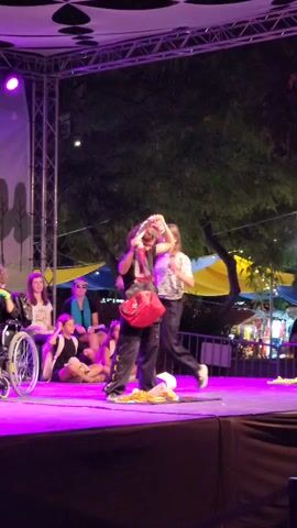 The video shows a performance on a stage. Two people are center stage. The person on the left is wearing a patterned shirt and dark pants and is facing left. The person on the right is wearing a black shirt with a red collar, dark pants, and has a red bag with a white cross on their shoulder. They are interacting with the red bag. The person on the right bends down and picks up the red bag, then places it on the stage floor. There are other people seated on the stage to the left, including one person in a wheelchair. The stage is lit with purple lights. The background shows trees and tents, suggesting an outdoor evening event.