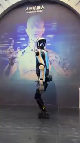 The video shows a humanoid robot standing on a dark gray floor in front of a backdrop with the words "HUMANOID ROBOT" in English and Chinese. The robot is primarily black and white with silver accents. It has a stylized head with a black visor and white casing. The body has a combination of black and white paneling, and the legs are black with thin, silver accents. The arms are mostly white with black details at the joints. The robot is in a bent posture, appearing to be holding its balance.  The backdrop shows a partial image of a humanoid figure and abstract digital patterns in the background. The area around the backdrop appears to be a display area.