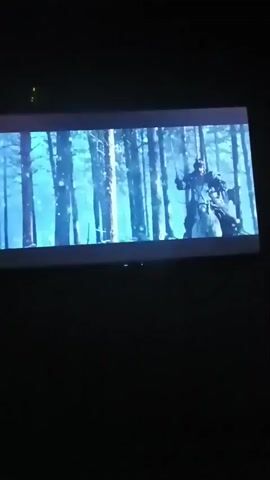 A video displays a cinematic scene on a screen, predominantly in blue and white tones. The scene appears to be set in a forest with numerous trees. In the first frame, a silhouette of a person is visible on the right side, facing left, with their arm extended. The subsequent frames reveal a person with a sword, dressed in armor, standing and then riding a horse. The person appears to be in motion, possibly in a battle or chase sequence. The final frame shows a close-up of a person wielding a glowing sword in a dark, possibly snowy environment. The video transitions through these visual elements rapidly, suggesting a trailer or action sequence.