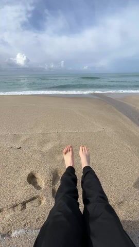 A first-person perspective shows two bare feet in the foreground, the person wearing black pants. Behind the feet is a sandy beach, with footprints in the sand. The ocean stretches into the distance with small waves breaking along the shoreline. The sky overhead is mostly cloudy with patches of blue.