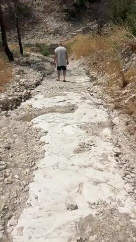 A person walks away from the camera on a dry, rocky path. The path is lined with dry grass and brush. In the distance, there are hills with sparse vegetation. The ground appears to be made up of small rocks and dirt, with some larger white rock formations visible. The person is wearing a white t-shirt and black shorts. The overall environment appears to be arid and natural.