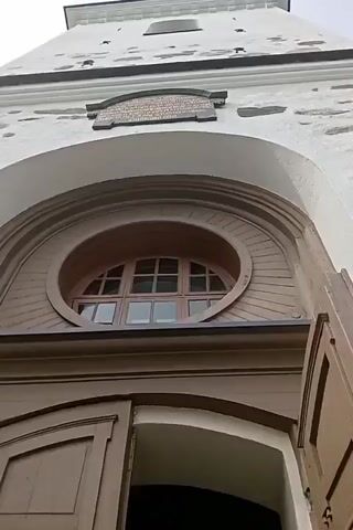 The video begins with an upward shot of the top of a large wooden door, with a round window above it. The camera then pans down and to the left, revealing the interior of a building. A red carpet runs down a hallway, leading to a set of large wooden doors. A chandelier hangs from the ceiling above the doors. The camera then moves to the right, showing a large screen mounted on the wall with text and images on it. The hallway continues to the right, with more doors visible in the distance. The video ends with a continued shot of the hallway and the screen.