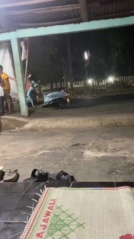 The video shows a dimly lit outdoor scene in Pegaden Tengah, Indonesia, likely at night. The foreground is dominated by a woven mat with the word "RAJAWALI" visible in red and green lettering. Several pairs of footwear, including sandals and slip-on shoes, are scattered on the tiled floor.

In the mid-ground, a light blue scooter is parked near a covered structure. A white wall with a red and white flag hangs on a pole behind it. To the left, two people are visible: an adult male wearing a long-sleeved shirt and shorts, and a young boy in a yellow t-shirt and shorts. The boy is actively engaged, leaning against a pillar and looking towards the street. The adult appears to be walking away from the camera.

The background reveals a street with sparse lighting from street lamps and passing vehicles, indicated by blurry streaks of light. A fence lines the side of the road. The overall atmosphere suggests a casual, perhaps residential, setting at night, with everyday activities taking place. The presence of footwear and the covered structure suggest a home or a local shop. The lighting indicates it is nighttime, and there are no obvious signs of extreme weather.