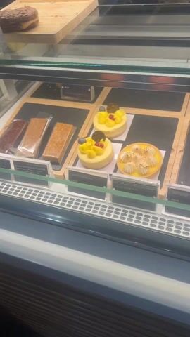 The camera pans across a refrigerated display case filled with various desserts and candies. On the top shelf, there are lemon cheesecakes, vegan lemon tarts, red velvet cake, carrot cake, and several chocolate and cream desserts. The second shelf contains chocolate treats in packages and an assortment of cakes and pastries. On the bottom shelf, there is a selection of chocolates, including squares, spheres, and heart-shaped pieces in red and yellow. The prices are indicated on small signs in front of the items.