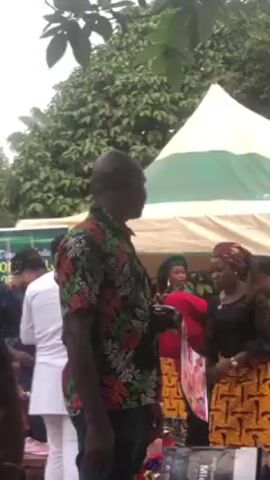 The video shows a crowd of people gathered outside under some trees and a white and green tent. A man in a floral shirt stands in the foreground, facing the crowd.  A man in a white outfit stands near the tent. Women in colorful outfits and head coverings are visible near a table under the tent. A small child stands among them. A person in a red shirt walks in front of the tent, towards the right. A laptop and a speaker are visible on a table under the tent. The music is playing loudly.