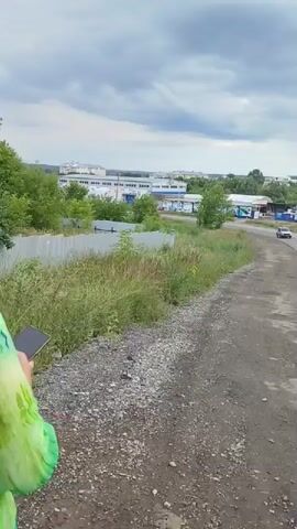 The video opens with a view of an overcast sky above a suburban or industrial area. In the foreground, a person wearing a green and tie-dye shirt is partially visible on the left, holding a dark object. The scene then pans to the right, showing a dirt road with tall grass and weeds growing along its edge. Further ahead, a paved road curves to the right, with several cars driving on it. On the left side of the paved road, there are low-lying buildings with light-colored facades and some green trees. On the right side of the paved road, more trees and vegetation are visible, along with a billboard. The overall environment appears to be a mixed landscape of nature and man-made structures, under a cloudy sky.