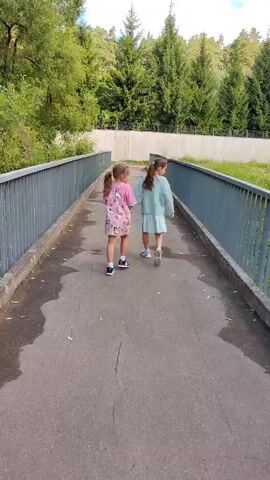 Two young girls walk away from the camera on a paved path bordered by metal railings on a bridge. The girl on the left wears a pink dress with a floral pattern and dark shoes. The girl on the right wears a light blue dress and dark shoes. They are walking towards a line of evergreen trees and deciduous trees. The sky above is blue with white clouds.