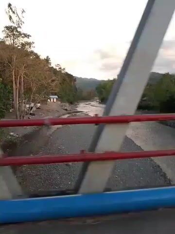 The video shows a view from a bridge looking downstream at a river flowing through a natural landscape. The riverbanks are lined with trees and vegetation, and in the distance, mountains are visible under a cloudy sky. There are some small buildings on the riverbanks, suggesting a rural or semi-rural settlement. The bridge itself has red and blue railings, and the camera appears to be moving, indicating the viewpoint is from a vehicle or on foot crossing the bridge. The light suggests it is either early morning or late afternoon. There are no people clearly visible in the video.