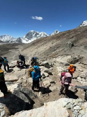 The video depicts a group of trekkers navigating a rocky, high-altitude trail under a clear blue sky, with magnificent snow-capped mountains forming a dramatic backdrop.

In the initial shot, several individuals wearing backpacks and outdoor gear are seen on a dirt and rock path. One person, with a backpack adorned with colorful prayer flags and a solar panel, stands on a rock, holding up a smartphone to capture the view. Other trekkers are seen resting, with some crouching or standing among the rocks.

The landscape is rugged and sparse, characterized by barren ground, loose rocks, and distant glaciers. Looming snow-covered peaks dominate the horizon, suggesting a remote and challenging environment. Pack animals, likely yaks or mules, carrying cargo are also visible, being guided by a person across the rocky terrain.

The scene conveys a sense of adventure and awe, with the trekkers seemingly pausing to take in the vast and beautiful mountain vista. The bright sunlight and absence of clouds indicate clear, sunny weather conditions. No visible text is present in the video.
