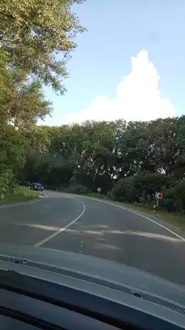 A dark-colored car drives on a winding road through a forest. The road has a white line marking the lane. Trees line both sides of the road. The sky is blue with some white clouds. The camera appears to be mounted inside another vehicle, looking forward.