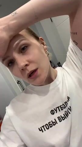 A person with light skin and blonde hair in a low ponytail is standing inside a room with white cabinets in the background. The person is wearing a white t-shirt with black Cyrillic text on it. The person's right arm is raised, with their hand resting on their forehead. The person's eyes are mostly closed, and they appear to be speaking or singing. The person has several small piercings in their left ear. The lighting in the room is bright, and the shot is close-up, focusing on the person's face and upper body.